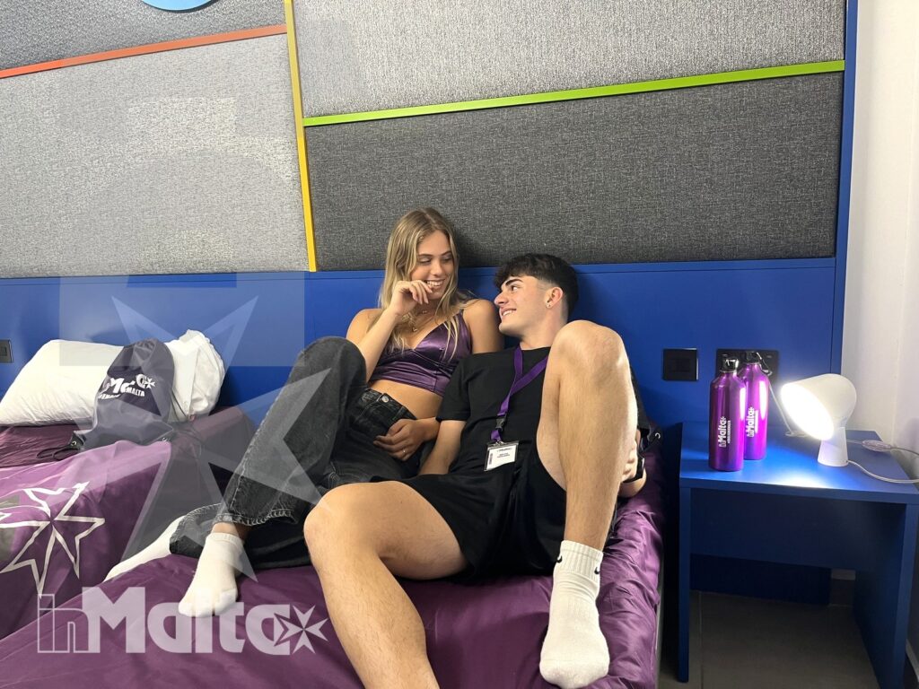 Giovani partecipanti condividono momenti quotidiani nell'alloggio a Malta Due studenti sorridono e parlano seduti su un letto in una camera condivisa colorata, tra zaini e bottiglie riutilizzabili.