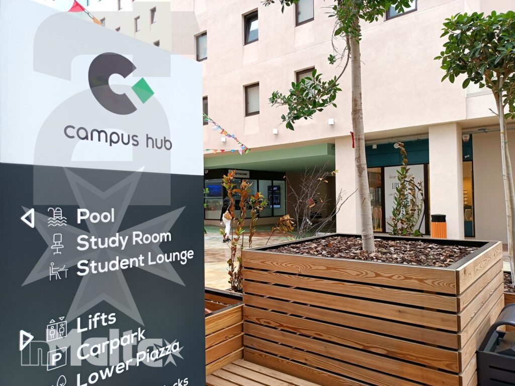 Cartello Campus Hub con indicazioni per piscina, sala studio, student lounge, ascensori e parcheggio nel cortile della struttura.