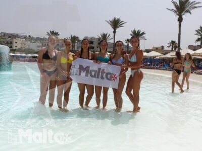 Sei studentesse sorridenti mostrano un cartello con la scritta Malta nell'acqua bassa di una piscina, tra palme e resort.