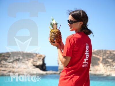 Studentessa sorridente tiene un ananas decorato davanti al mare turchese e alle scogliere calcaree della costa maltese.