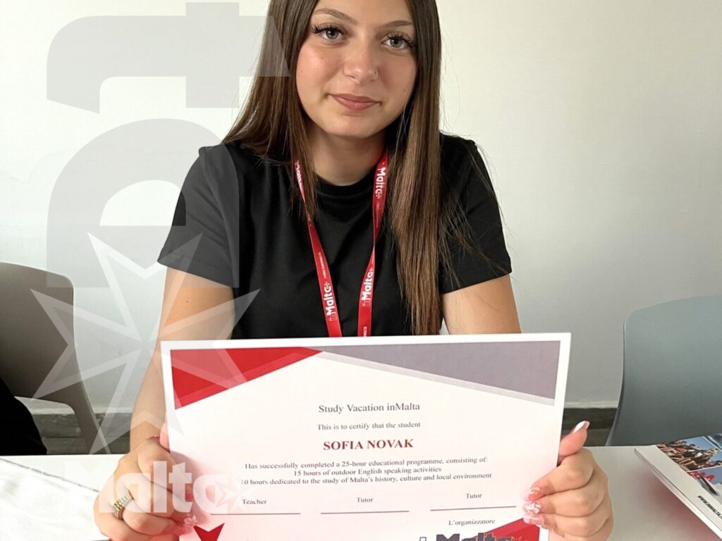 Studentessa sorridente mostra un certificato personalizzato alla fotocamera, al termine di un'esperienza formativa con il brand inMalta.