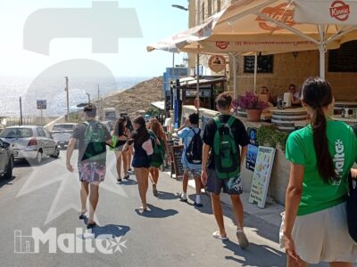 Studenti con zaini camminano lungo una strada costiera in discesa verso il mare, tra edifici in pietra maltese durante l'escursione.