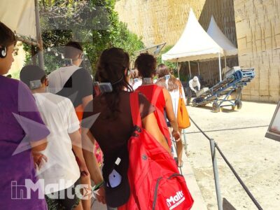 Studenti con zaini e borse camminano in fila tra mura storiche, transenne e una tensostruttura durante una visita organizzata a Malta.