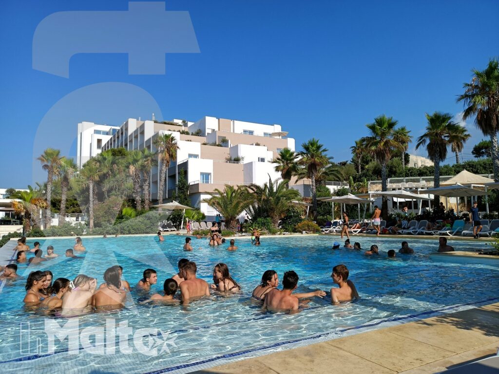 Studenti inMalta nella piscina del resort Studenti e altri ospiti si ritrovano nella piscina del resort, con hotel, lettini, ombrelloni e palme sullo sfondo.
