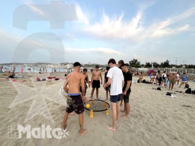 Giovani partecipanti giocano attorno a una rete bassa sulla spiaggia, con il mare e altri bagnanti sullo sfondo.