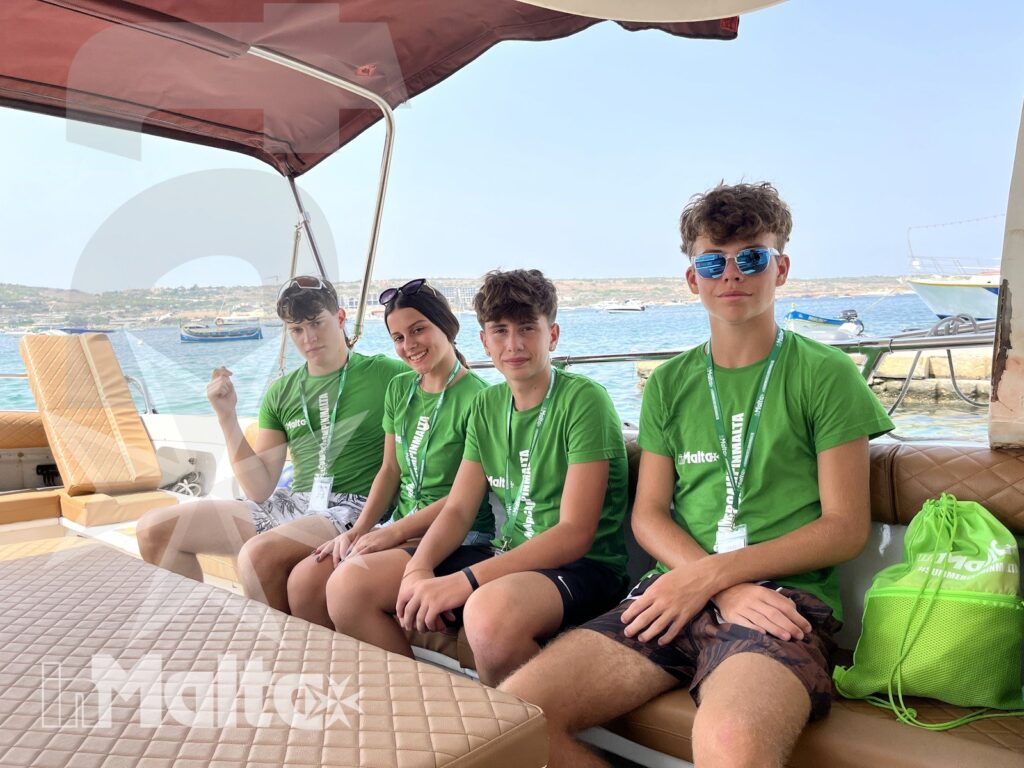 Quattro studenti sorridono seduti insieme sulla barca, con il panorama costiero di Comino e il mare alle spalle.