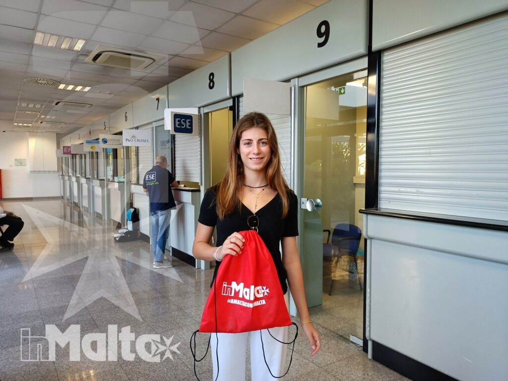 Studentessa inMalta alla reception della scuola Studentessa sorridente con borsa rossa davanti alla reception della scuola, in un corridoio frequentato da altri studenti in vacanza studio.