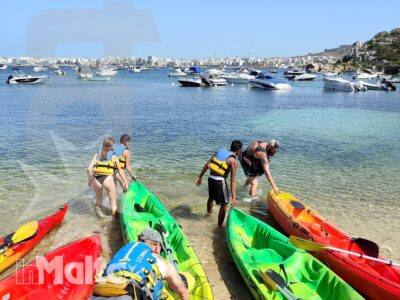 Un gruppo di studenti spinge kayak colorati in una baia maltese con barche e costa abitata sullo sfondo, in un'attività inMalta.