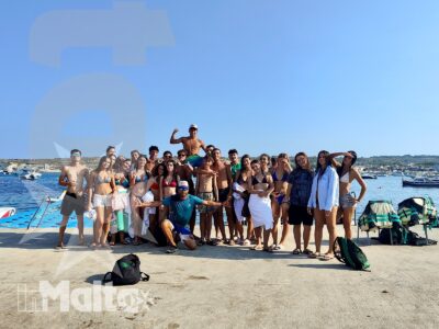 Gruppo numeroso di studenti posa su un molo tra barche e costa, durante un'attività estiva sul mare a Malta.