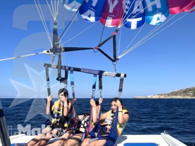 Due giovani sorridono sul bordo di un catamarano mentre praticano parasailing, con mare aperto e costa rocciosa alle spalle.