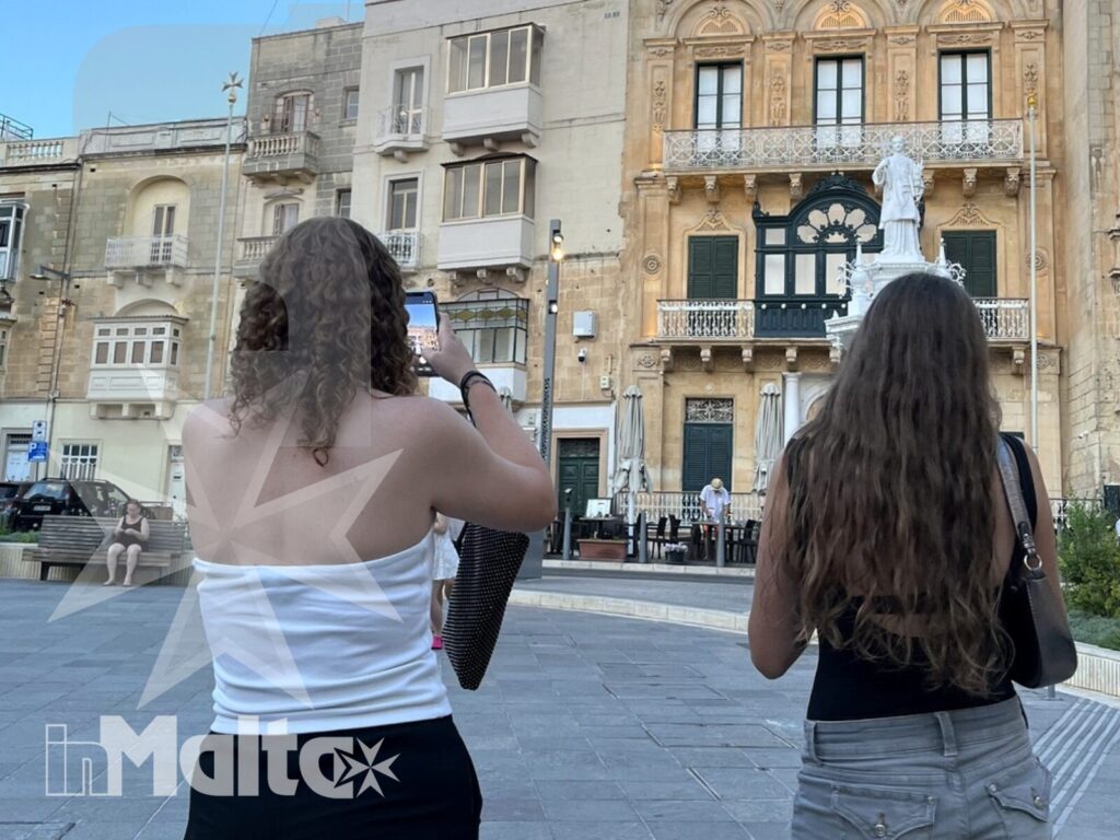 Studentesse alla scoperta delle piazze storiche maltesi Due studentesse osservano edifici storici decorati in una piazza maltese durante una visita culturale alle Tre Città.
