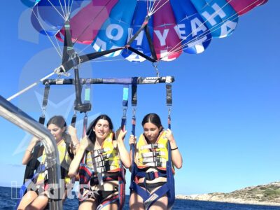 Tre giovani sono sollevati sopra il mare dalla barca, sotto un parasail decorato e con una costa rocciosa sullo sfondo.