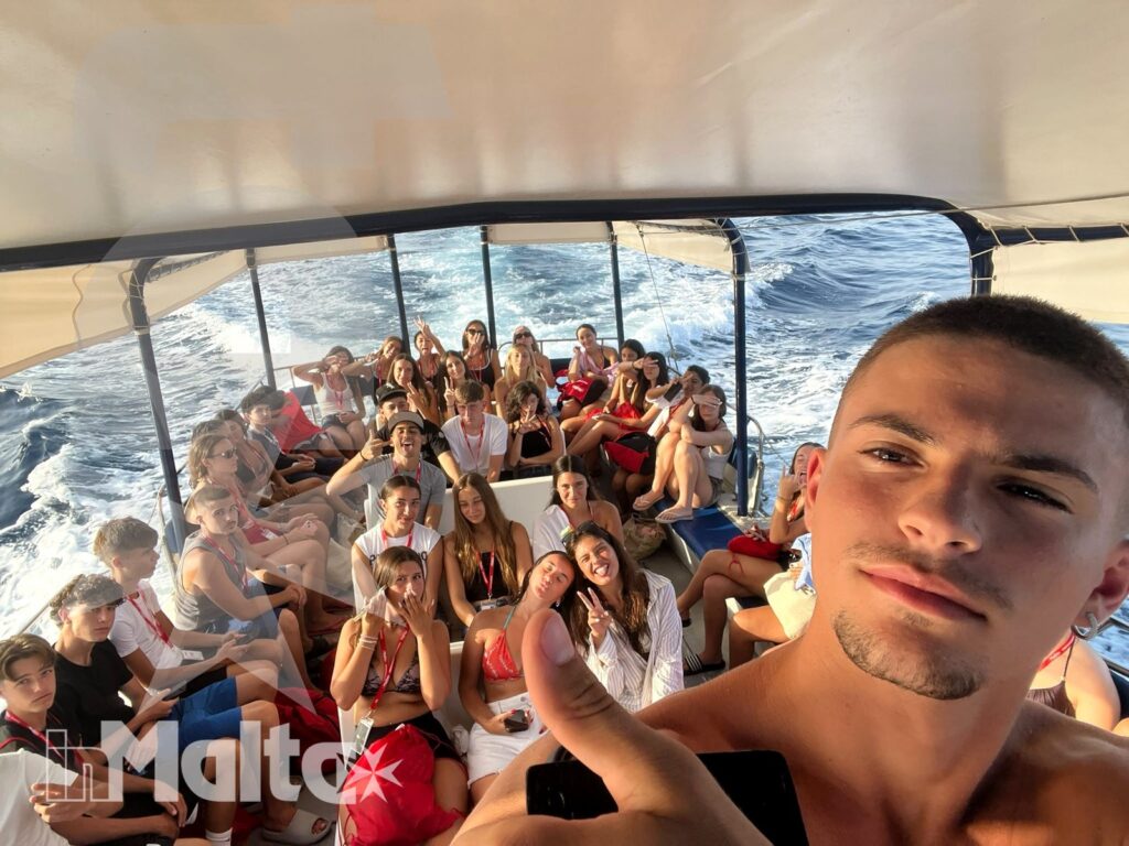 Studente in primo piano fa il pollice in su, mentre il gruppo siede a bordo durante la navigazione e il mare appare sullo sfondo.