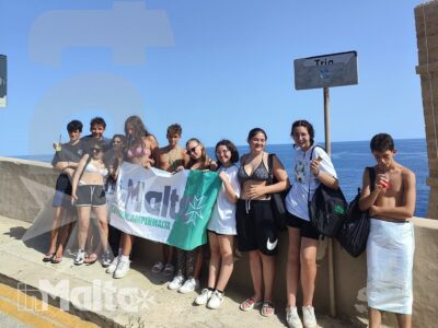 Gruppo di studenti posa informalmente con uno striscione sul lungomare maltese, mentre mare e cielo sereno incorniciano l'esperienza di viaggio.