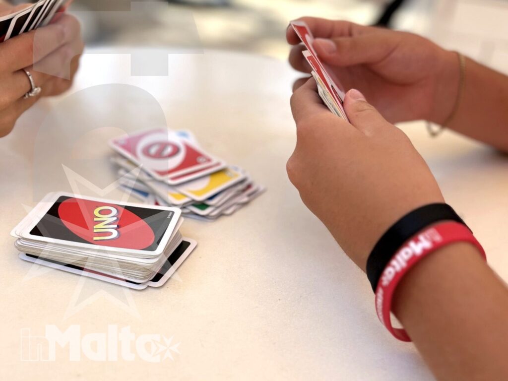 Dettaglio delle mani di più studenti impegnati in una partita a UNO all'aperto, con carte e braccialetti identificativi.
