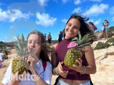 Due studentesse sorridenti tengono bevande servite in ananas sul litorale roccioso, condividendo un momento conviviale del soggiorno studio.