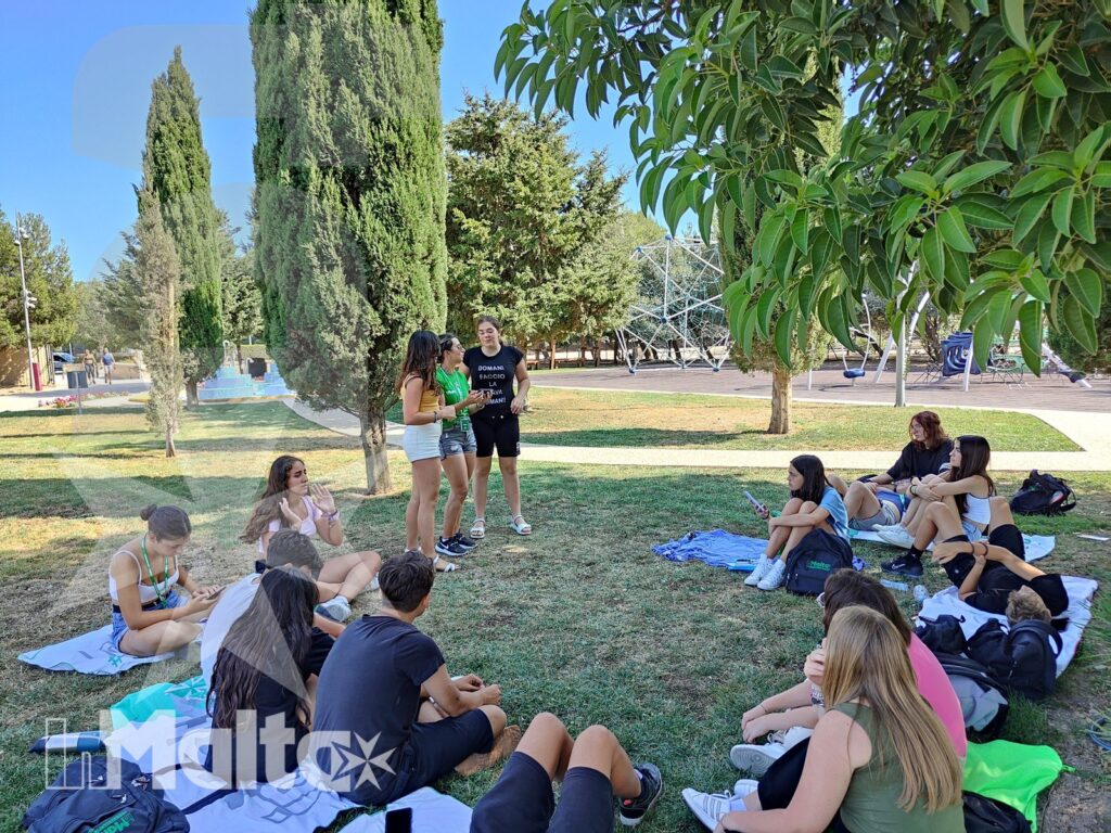 Studenti conversano in piccoli gruppi sul prato, accanto a teli, zaini e una piscina, durante una pausa nell'area ricreativa.