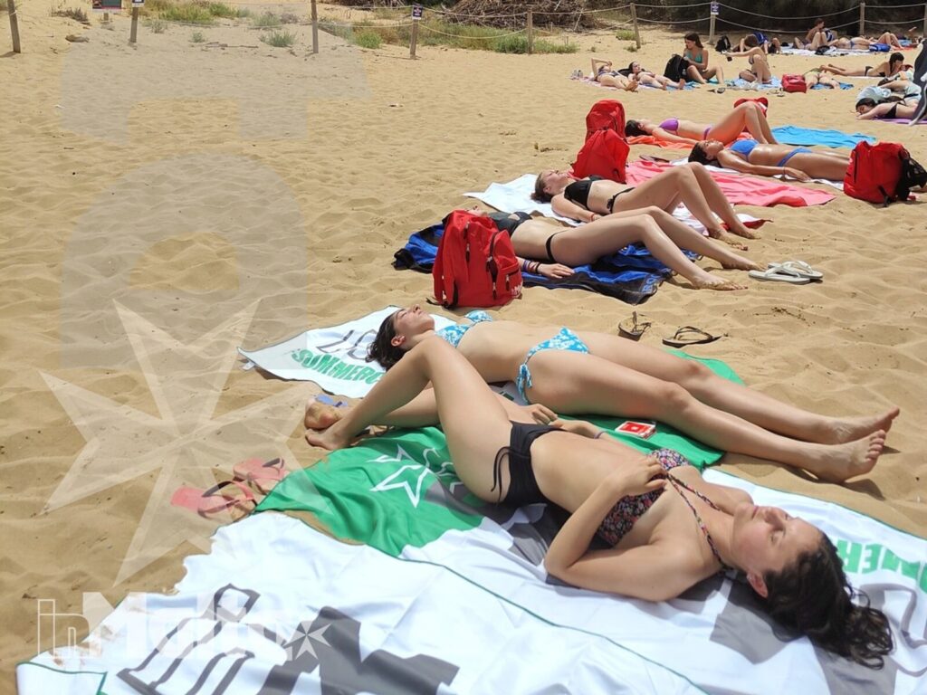 Gruppo di studenti si rilassa sulla sabbia tra asciugamani colorati e vegetazione mediterranea, durante una giornata balneare a Golden Bay.