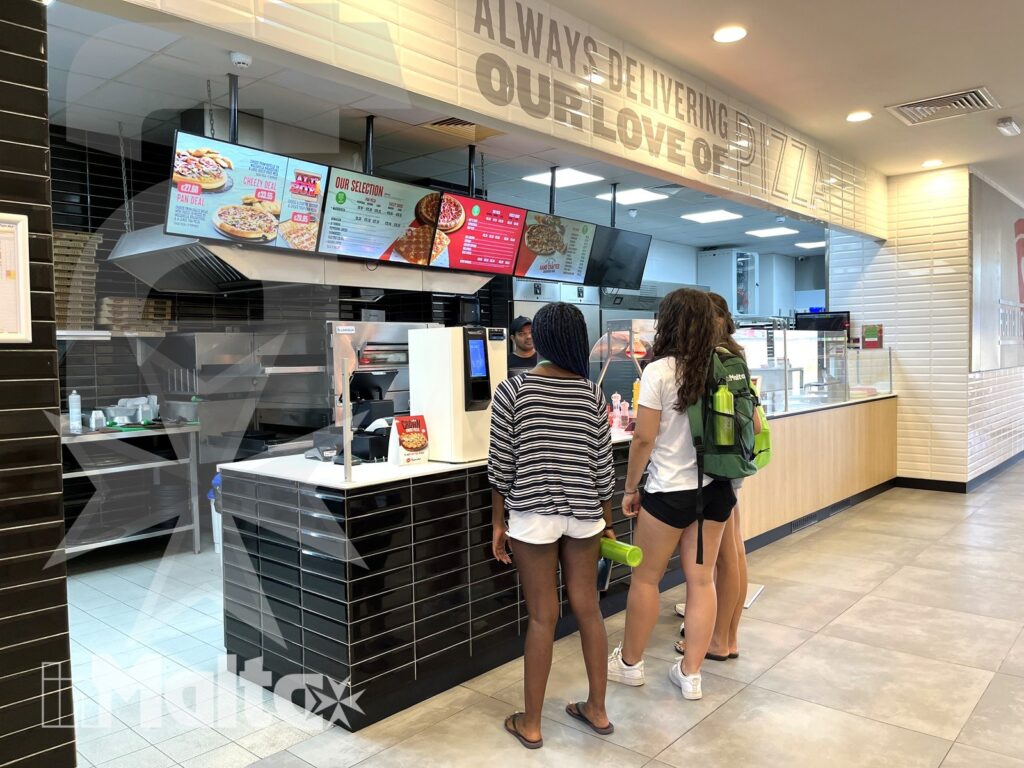 Due studentesse ordinano al banco di un ristorante con menu digitali e cucina a vista.