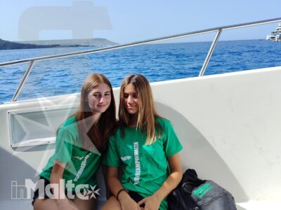 Due studentesse sorridono sedute su una barca in navigazione, con uno zaino e la costa maltese visibili sullo sfondo.