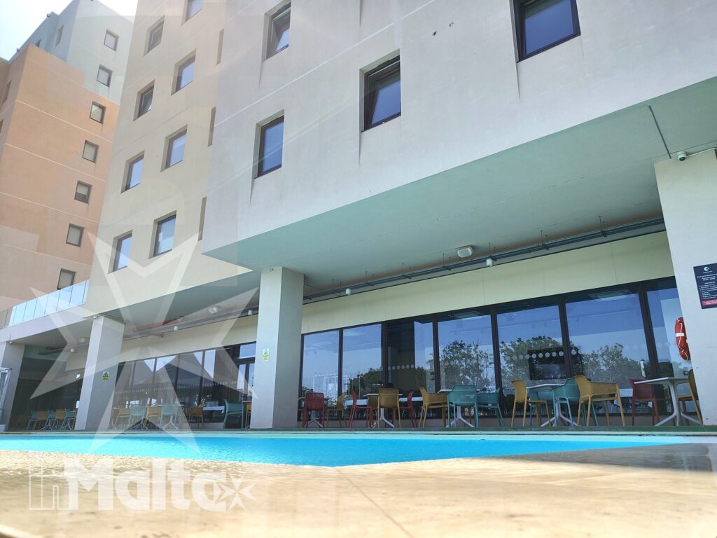 Piscina in primo piano davanti alla facciata moderna del Campus Hub, con area ristorante vetrata e tavoli all'aperto.