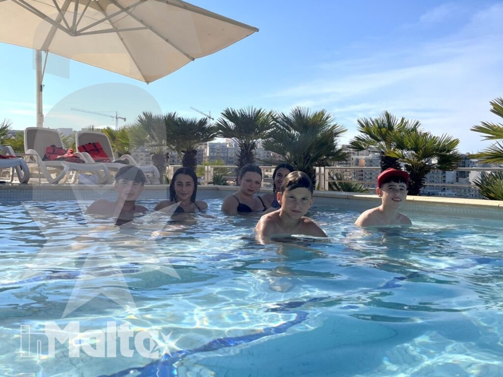 Gruppo di studenti immersi nella piscina soleggiata, tra palme e ombrellone, durante un soggiorno studio a Malta.