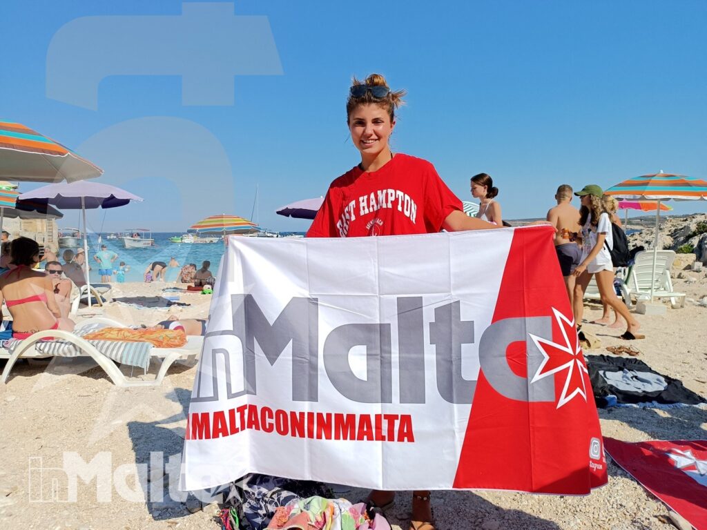 Studentessa inMalta mostra la bandiera sulla spiaggia Studentessa sorridente mostra una bandiera del programma sulla spiaggia, tra ombrelloni colorati e il mare sullo sfondo.