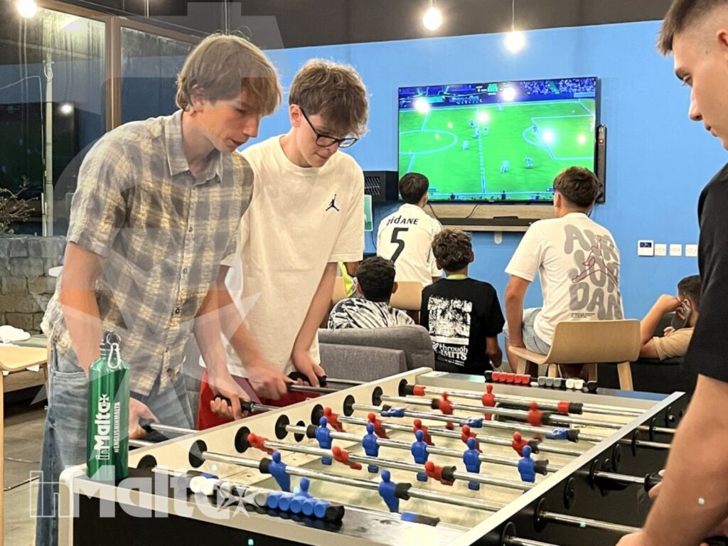 Studenti giocano a calcio balilla davanti a uno schermo con una partita di calcio, nella vivace sala comune di un alloggio inMalta.