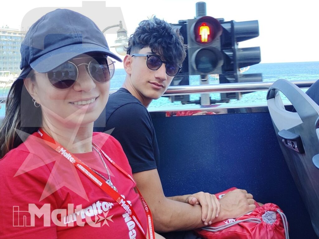 Accompagnatrice e studente siedono sull'autobus scoperto durante un'escursione a Malta, con il mare visibile sullo sfondo.