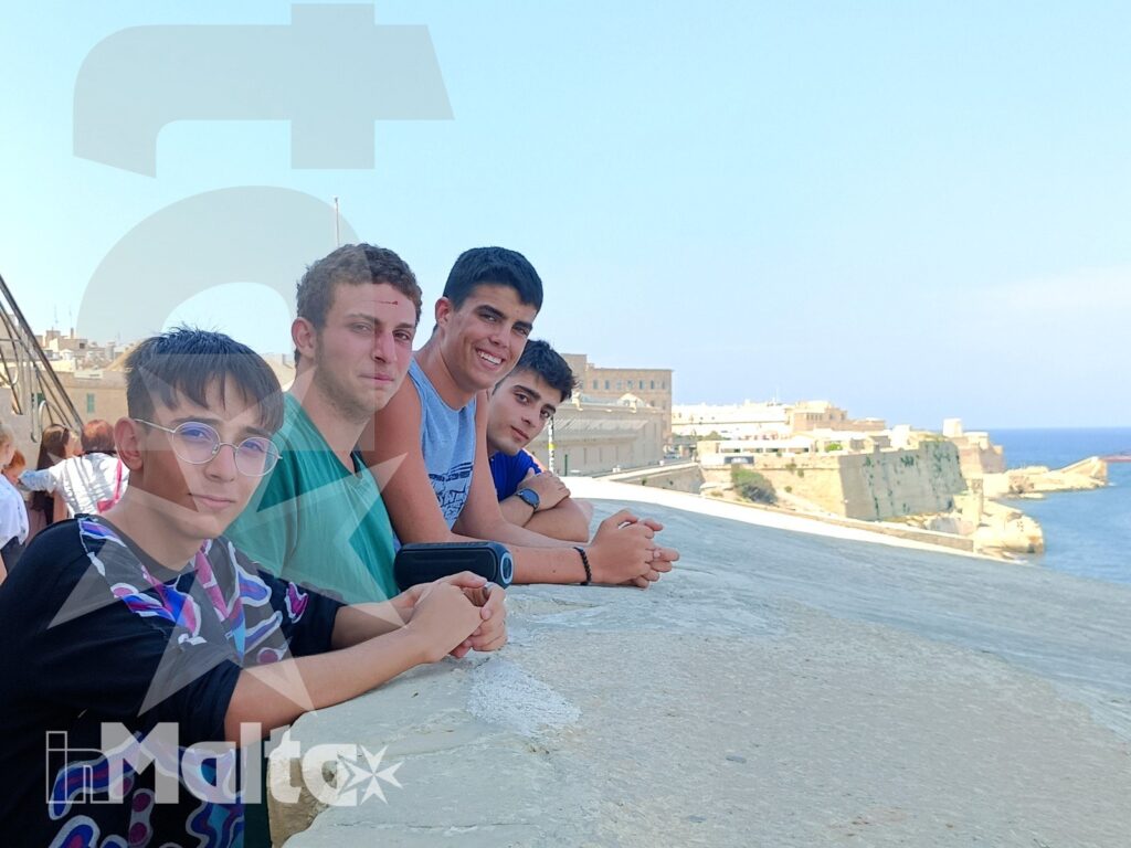 Studenti davanti alle fortificazioni storiche sul mare di Malta Quattro studenti sorridenti si affacciano sul mare accanto a fortificazioni storiche durante una visita culturale a Malta.