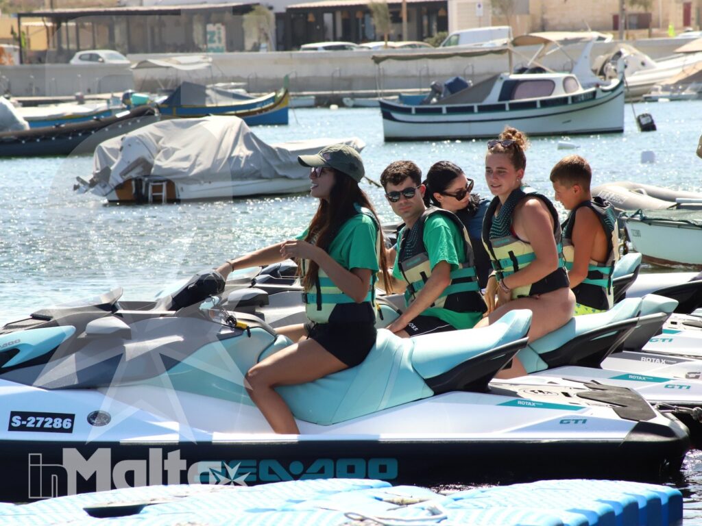 Un gruppo di giovani con giubbotti salvagente è seduto su moto d'acqua ferme in un porto turistico pieno di imbarcazioni.