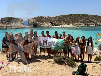 Gruppo numeroso di studenti posa sulla spiaggia davanti a una baia turchese e alle scogliere, durante l'escursione a Comino.