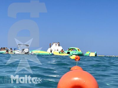 Giovani nuotano e sostano su strutture galleggianti, tra boe arancioni e gommoni, durante un'attività acquatica in mare.