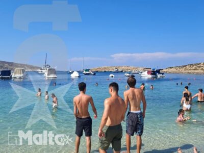 Tre giovani entrano nell'acqua turchese di una baia maltese, circondati da bagnanti, barche e costa rocciosa.