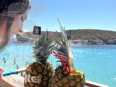 Due cocktail serviti in ananas spiccano su una terrazza affacciata sulla baia turchese, con una studentessa parzialmente visibile.