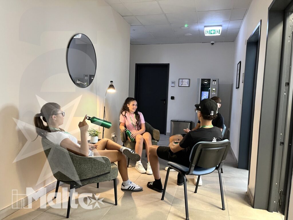 Quattro giovani studenti conversano seduti in cerchio nell'area comune interna, accanto a poltrone, distributore automatico e pianta.