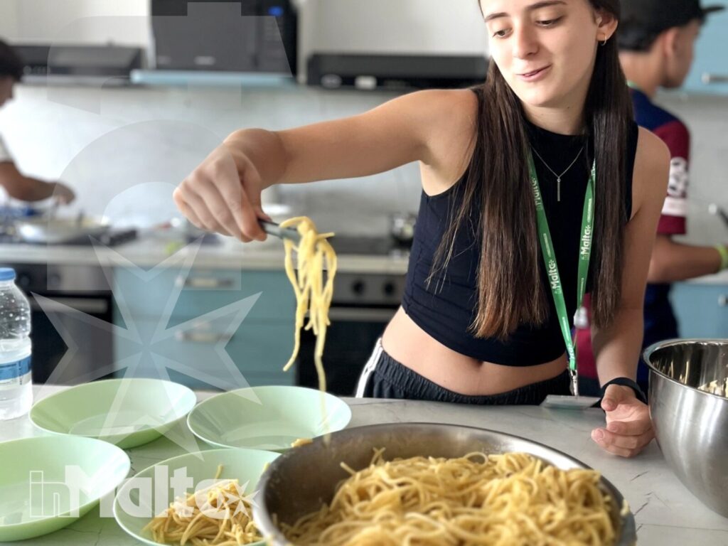 Studentessa solleva spaghetti dalle ciotole nella cucina comune, servendo la pasta durante un'attività culinaria con altri partecipanti.