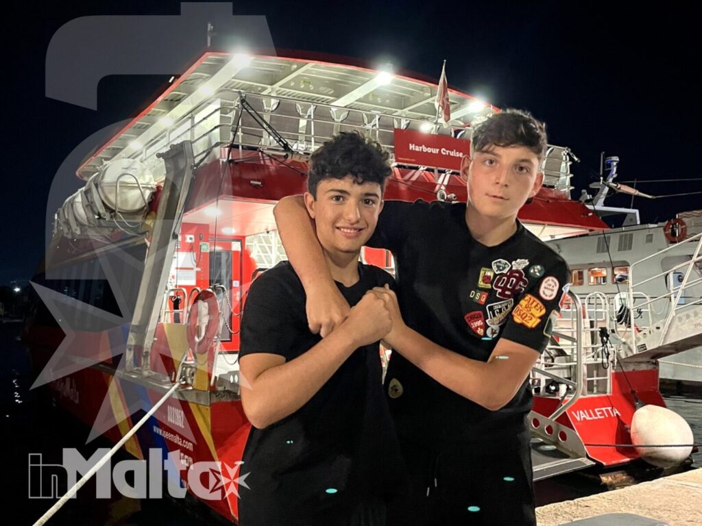 Due studenti sorridono davanti a un autobus turistico scoperto illuminato durante una visita serale a Malta.