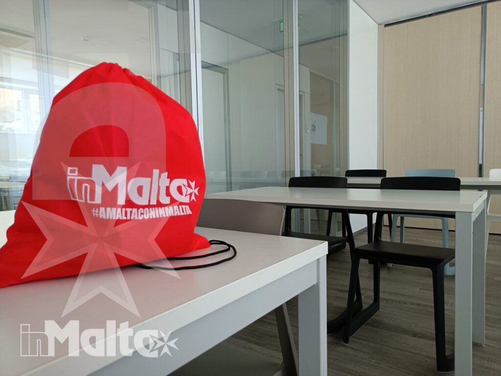Borsa rossa con logo inMalta appoggiata su un banco, davanti a un'aula moderna vuota con pareti vetrate.