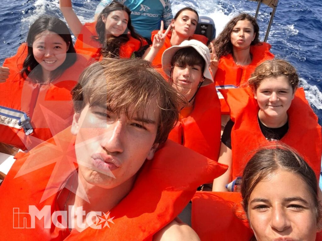 Gruppo di studenti con giubbotti salvagente arancioni posa per un selfie a bordo, salutando durante un'attività nautica in mare aperto.