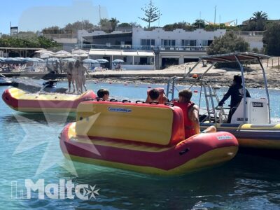 Giovani partecipanti a bordo di gommoni colorati prendono parte a un'attività organizzata, con barca di supporto e strutture balneari sulla costa.