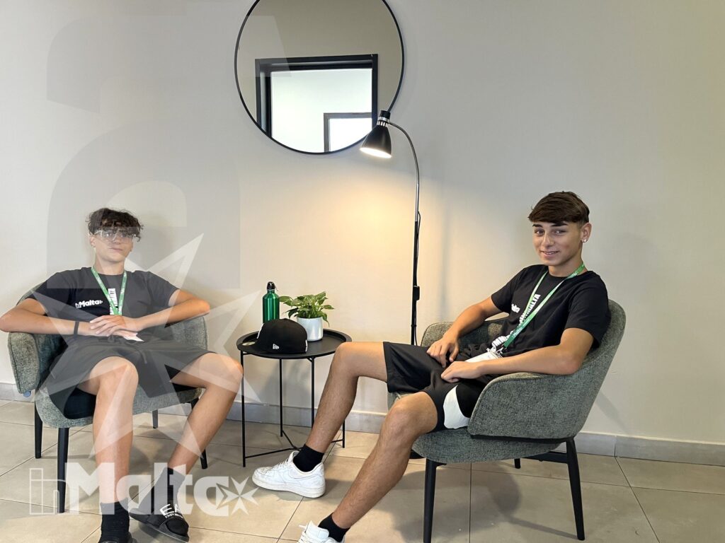 Area comune per studenti durante una vacanza studio Due studenti rilassati siedono su poltrone in una luminosa area lounge, con tavolino, specchio e pianta decorativa.