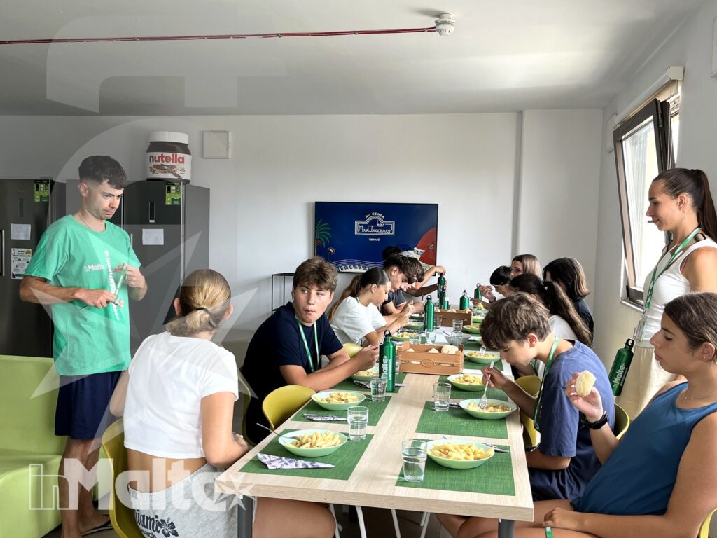 Gruppo internazionale di studenti pranza insieme attorno a una tavola apparecchiata, in un ambiente luminoso e informale del soggiorno studio.