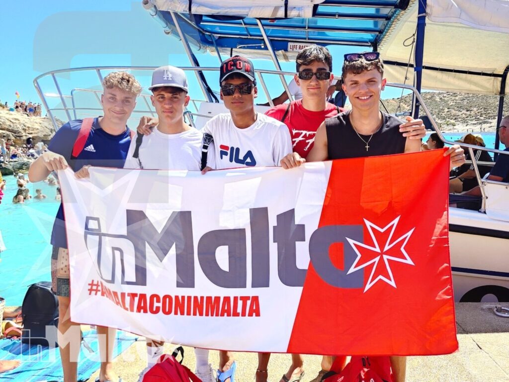 Studenti inMalta sulla barca verso la Blue Lagoon Studenti sorridenti mostrano una grande bandiera di Malta sul ponte della barca durante l'escursione alla Blue Lagoon.