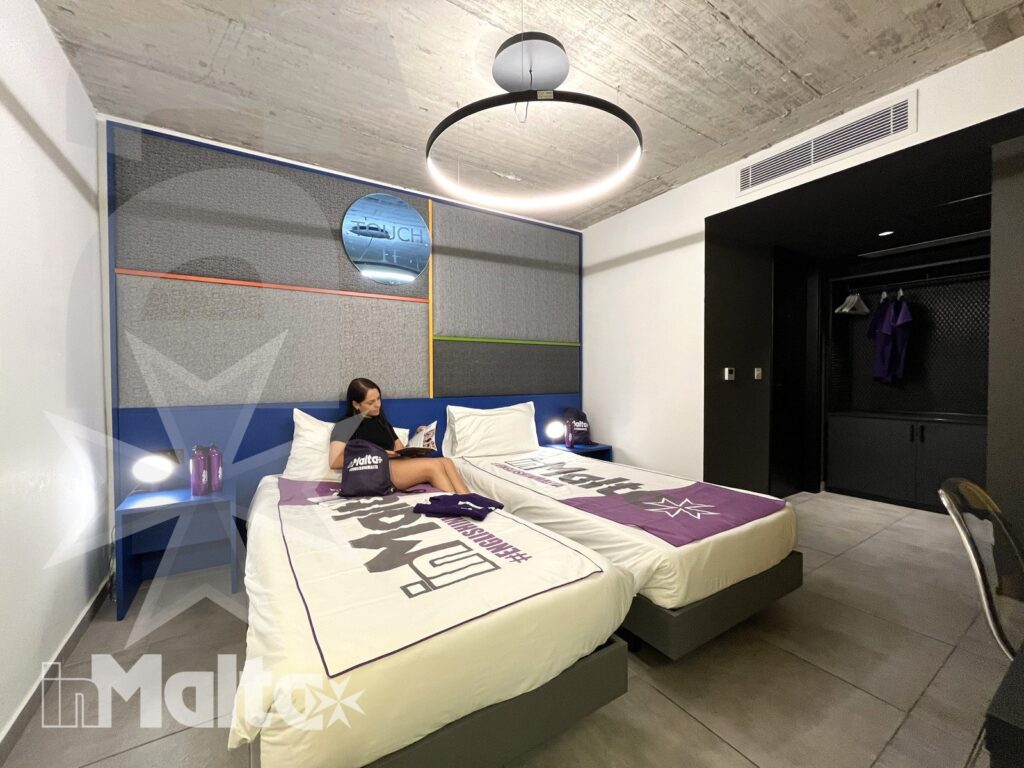 Sistemazione con letti singoli per partecipanti a Malta Camera contemporanea con due letti singoli, biancheria personalizzata e giovane ospite seduta mentre utilizza un dispositivo.