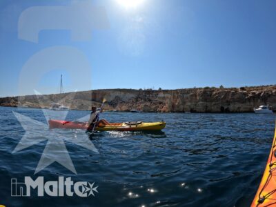 Una persona pagaia su un kayak colorato nelle acque limpide di Malta, con scogliere costiere illuminate dal sole sullo sfondo.