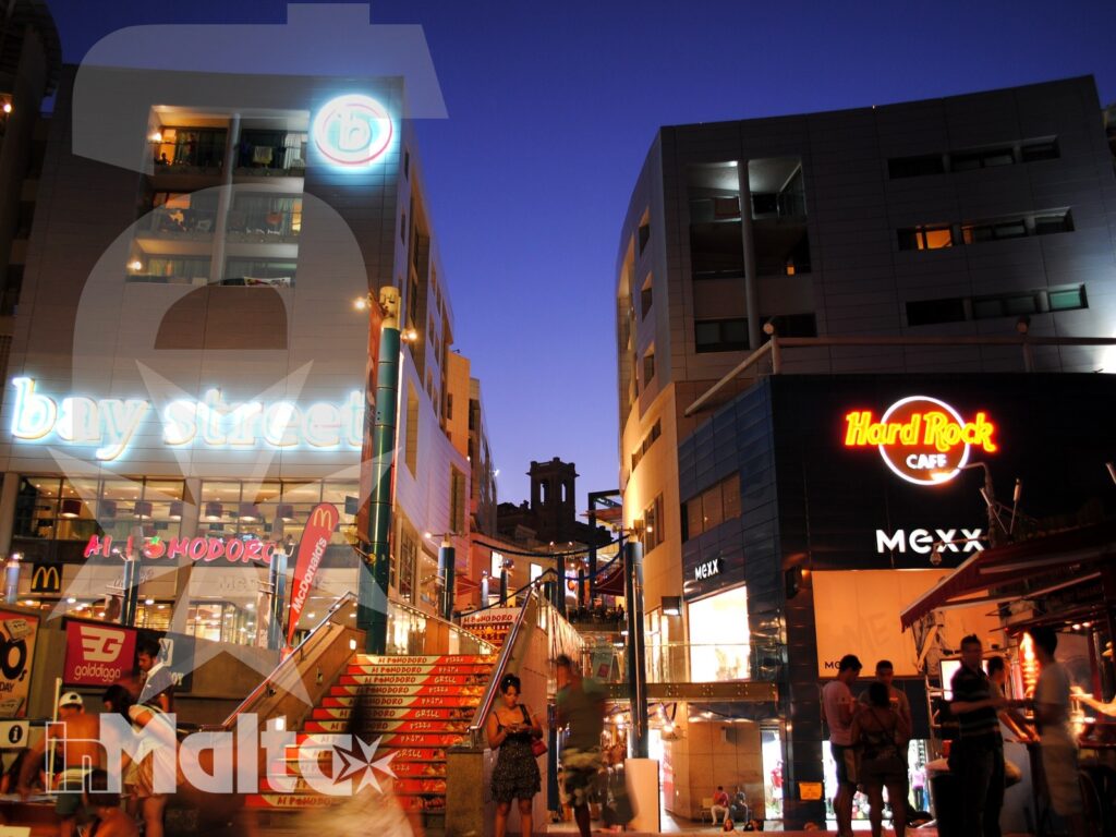 Strada commerciale notturna con scale illuminate, locali, insegne di Bay Street e Hard Rock Cafe, animata da passanti.