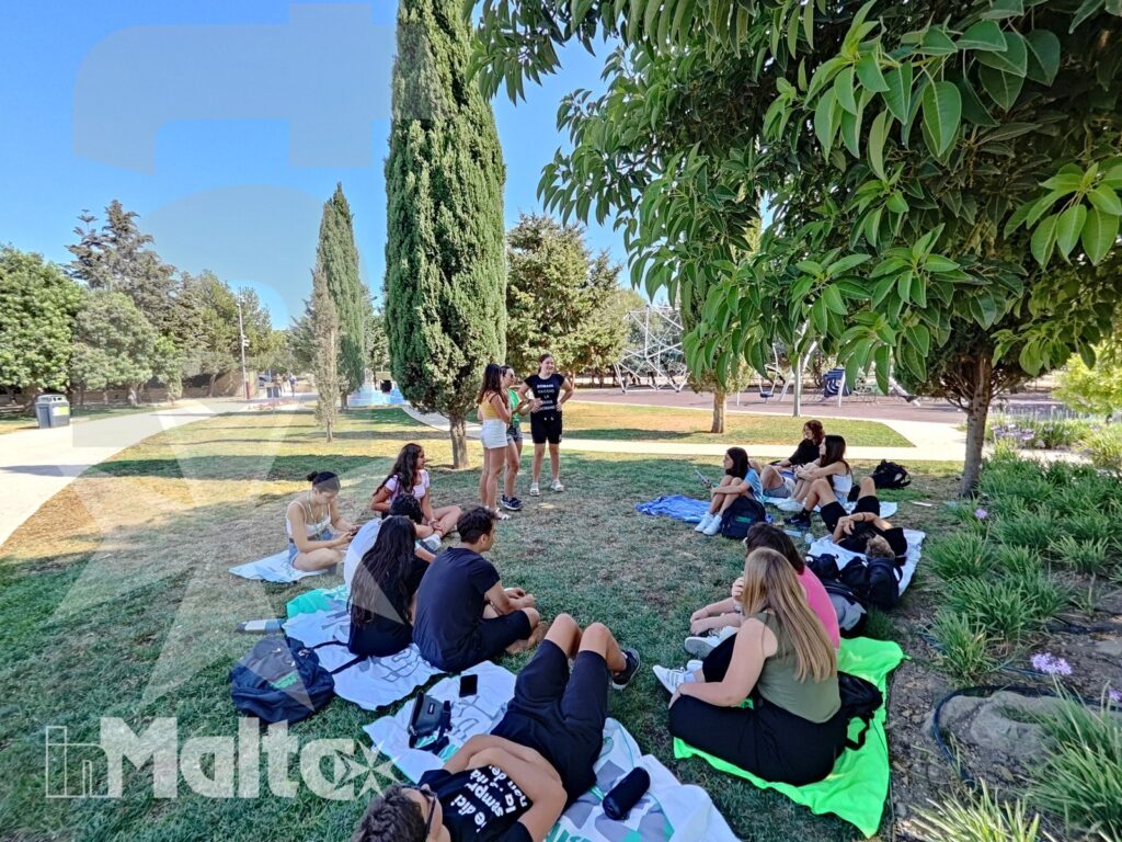 Studenti internazionali conversano sul prato ombreggiato di un giardino mediterraneo, durante una pausa dall'attività a Malta.