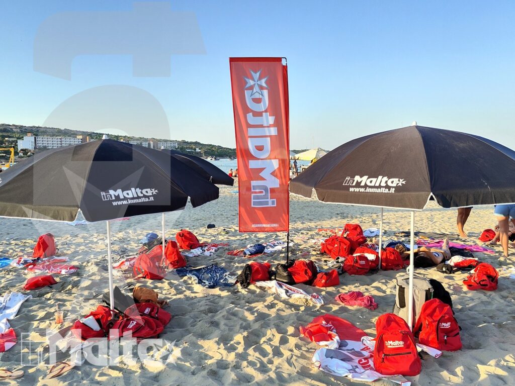 Allestimento per studenti sulla spiaggia di Mellieha Bay Ombrelloni, zaini rossi e bandiere organizzati sulla sabbia, con la costa di Mellieha Bay sullo sfondo per l'attività inMalta.