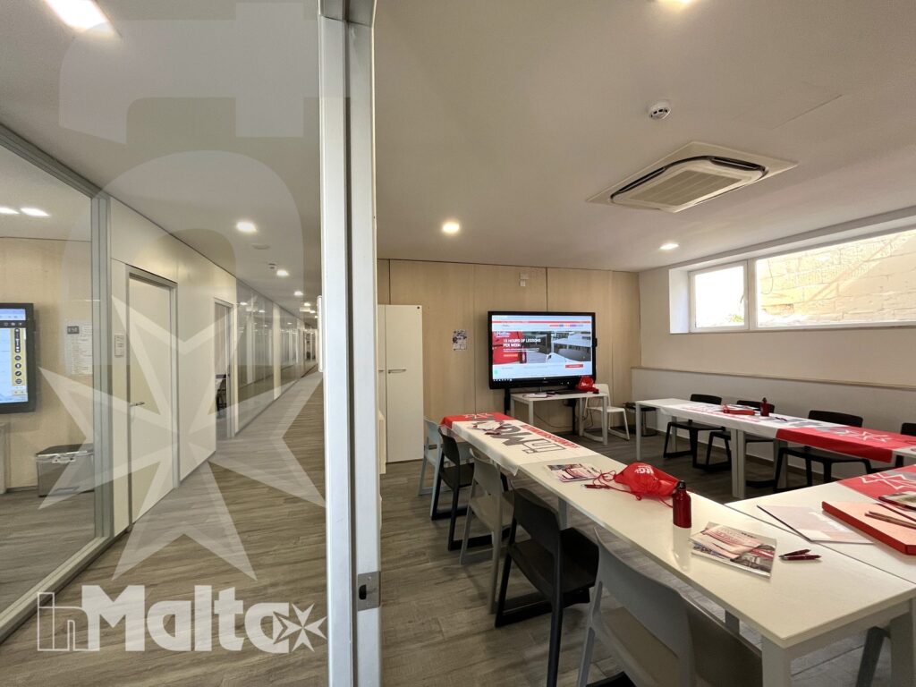 Aula scolastica luminosa con tavoli a isole, dispense, monitor acceso e corridoio vetrato, pronta ad accogliere gli studenti.